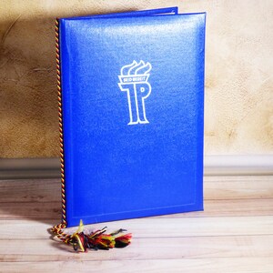 May include: A blue book with a white emblem and the text "SEID BEREIT" and "TP". The book has a decorative spine with red, yellow, and black threads. A tassel with similar colors lies at the bottom.