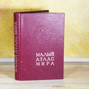 May include: A maroon hardcover book titled "МАЛЫЙ АТЛАС МИРА" in white Cyrillic lettering on the front cover and spine. The front cover features a debossed circular globe graphic above the text. The book has a textured finish and stands upright.