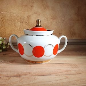 May include: White ceramic sugar bowl with a lid, featuring a vibrant orange and gold design. The bowl has two handles and a gold-coloured knob on the lid. The design includes large orange circles and gold sunburst patterns. A classic piece for serving sugar.