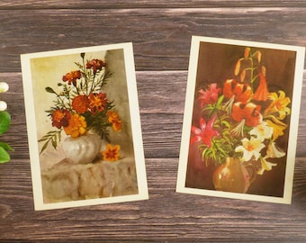 Puede incluir: Dos láminas rectangulares con bodegones florales. La lámina de la izquierda muestra caléndulas naranjas y rojas en un jarrón blanco. La de la derecha presenta un ramo de lirios rojos, naranjas y blancos en una jarra de cerámica marrón.