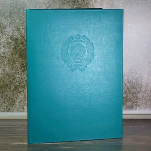May include: A teal-colored book with a textured cover. The cover features a circular embossed emblem in the center. The book is closed and appears to be in good condition. The background is a neutral color.