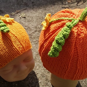 Könnte beinhalten: Zwei gestrickte Kürbis-Mützen mit grünen Stielen und gelben Blumenakzenten. Die Mützen sind orange und haben eine gerippte Textur.