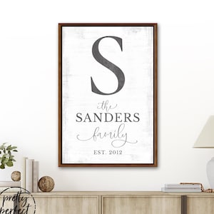 Puede incluir: Un letrero familiar enmarcado con un borde marrón oscuro. El letrero presenta una gran letra gris "S" en la parte superior, seguida de "the SANDERS family" en texto gris y "EST. 2012" en la parte inferior. El fondo es blanco con una textura envejecida.