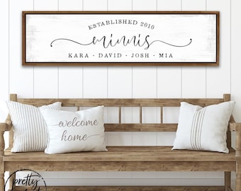 Può includere: Un cartello da parete rettangolare con sfondo in legno bianco e cornice marrone scuro. Il testo recita ESTABLISHED 2010, minnis, e i nomi KARA, DAVID, JOSH, MIA. Il cartello è esposto sopra una panca in legno con tre cuscini decorativi.