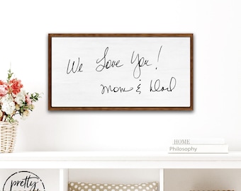 Puede incluir: Un letrero rectangular de madera con un marco marrón oscuro y fondo blanco. El letrero presenta un texto negro escrito a mano que dice We Love You! mom & dad. El letrero está montado en una pared lisa sobre un estante blanco con objetos.