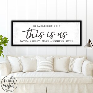 May include: A black framed wooden sign with the text "this is us" in a cursive font. Below the text are the names David, Ashley, Noah, Jennifer, and Kyle. The sign is on a white shiplap wall above a cream-colored sofa.