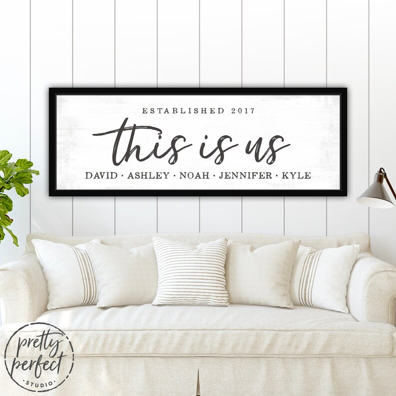 May include: A black framed wooden sign with the text "this is us" in a cursive font. Below the text are the names David, Ashley, Noah, Jennifer, and Kyle. The sign is on a white shiplap wall above a cream-colored sofa.