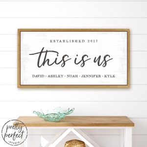 May include: A framed wooden sign with the words "this is us" in black script. Below the text are the names David, Ashley, Noah, Jennifer, and Kyle. The sign is on a white background and reads "Established 2017".