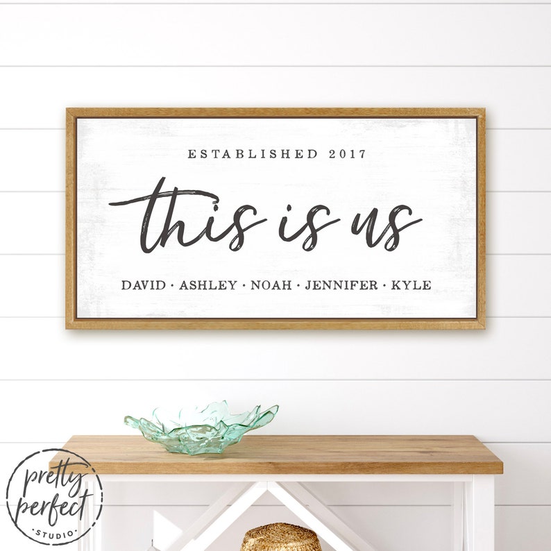 May include: A framed wooden sign with the words "this is us" in black script. Below the text are the names David, Ashley, Noah, Jennifer, and Kyle. The sign is on a white background and reads "Established 2017".