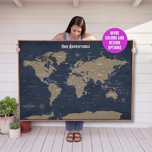 May include: A framed world map with a navy blue background and gold-colored continents. The map is labeled "Our Adventures" and includes the family name "Davis Family". A pink circle with the text "MORE COLORS AND DESIGN OPTIONS" is in the upper right corner.