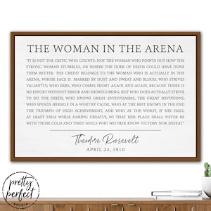 May include: A printable quote by Theodore Roosevelt about the woman in the arena. The quote is printed in black text on a white background with a brown wooden frame. The quote reads, "It is not the critic who counts: not the woman who points out how the strong woman stumbles, or where the doer of deeds could have done them better. The credit belongs to the woman who is actually in the arena, whose face is marred by dust and sweat and blood; who strives valiantly; who errs, who comes short again and again, because there is no effort without error and shortcoming; but who does actually strive to do the deeds; who knows great enthusiasms, the great devotions; who spends herself in a worthy cause, who at the best knows in the end the triumph of high achievement, and who at the worst, if she fails, at least fails while daring greatly, so that her place shall never be with those cold and timid souls who neither know victory nor defeat." The quote is signed "Theodore Roosevelt" and dated "April 23, 1910".