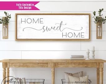 May include: A rectangular wall sign with a light wood frame and a white background featuring the gray text HOME sweet HOME. The sign is centered above a wooden console table with two wire baskets and two wall-mounted glass vases with greenery.