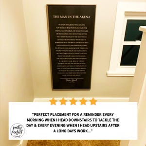 Puede incluir: Un letrero de pared rectangular con fondo negro y texto blanco que presenta la cita The Man in the Arena de Theodore Roosevelt. El letrero est&aacute; enmarcado con un borde oscuro fino y est&aacute; montado en una pared clara cerca de un marco de ventana.