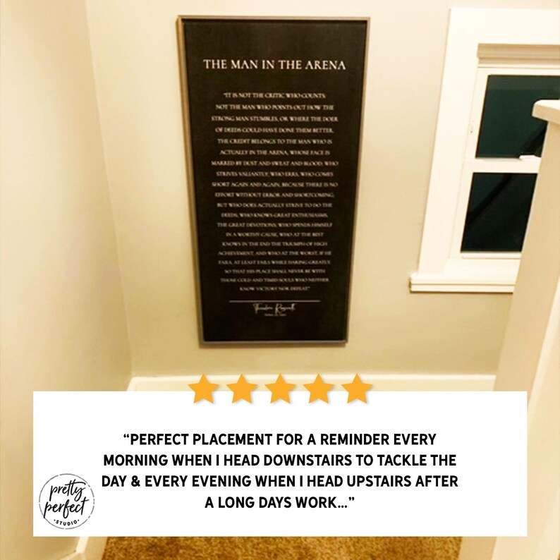 Puede incluir: Un letrero de pared rectangular con fondo negro y texto blanco que presenta la cita The Man in the Arena de Theodore Roosevelt. El letrero est&aacute; enmarcado con un borde oscuro fino y est&aacute; montado en una pared clara cerca de un marco de ventana.