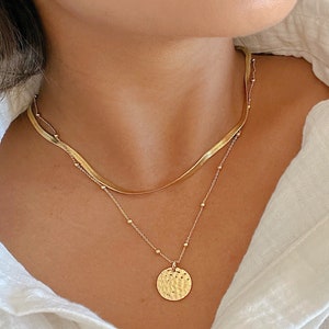 May include: Gold-toned jewelry set featuring a layered necklace and hoop earrings. The necklace includes a snake chain and a delicate chain with a textured pendant. The hoop earrings are adorned with small, sparkling accents. The jewelry is worn with a white collared shirt.