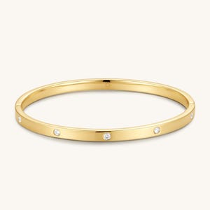 Może przedstawiać: Polerowana złota bransoletka typu bangle o smukłym, okrągłym kształcie. Powierzchnia metalu jest gładka i odblaskowa, ozdobiona kilkoma małymi, przezroczystymi, okrągłymi kamieniami osadzonymi w regularnych odstępach na zewnętrznym obwodzie.