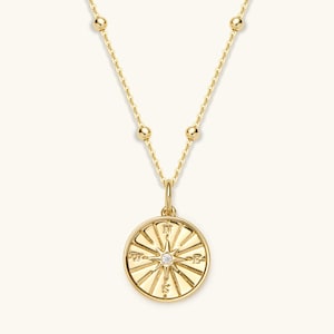 May include: A gold necklace featuring a compass pendant. The pendant has a small diamond at its centre and is surrounded by sunburst rays. The chain has small, evenly spaced gold beads.