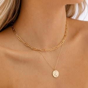 May include: A layered gold necklace set consisting of two distinct chains. The upper chain features elongated rectangular links, while the lower chain is a delicate cable style with a round, embossed coin pendant hanging from the center.