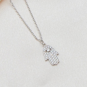 Könnte beinhalten: Eine silberfarbene Halskette mit einem Hamsa-Hand-Anhänger. Der Anhänger ist mit kleinen, klaren Kristallen und einem Augenmotiv besetzt. Die Halskette hängt an einer feinen Kette vor einem cremefarbenen Stoff.
