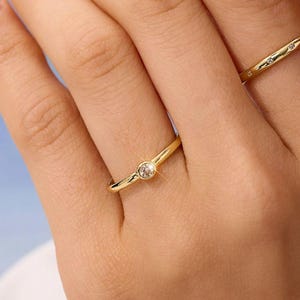 Op de afbeelding: Twee gouden ringen worden op een hand getoond. Eén ring heeft een enkele, heldere edelsteen. De andere ring heeft kleine, heldere edelstenen. De ringen staan tegen een zachte, vage achtergrond.