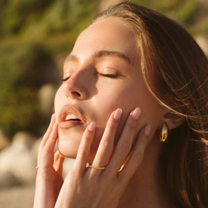 May include: A woman with long brown hair is touching her face with both hands. She is wearing a gold ring on her left ring finger and a gold hoop earring in her right ear. Her eyes are closed and she appears to be relaxed.