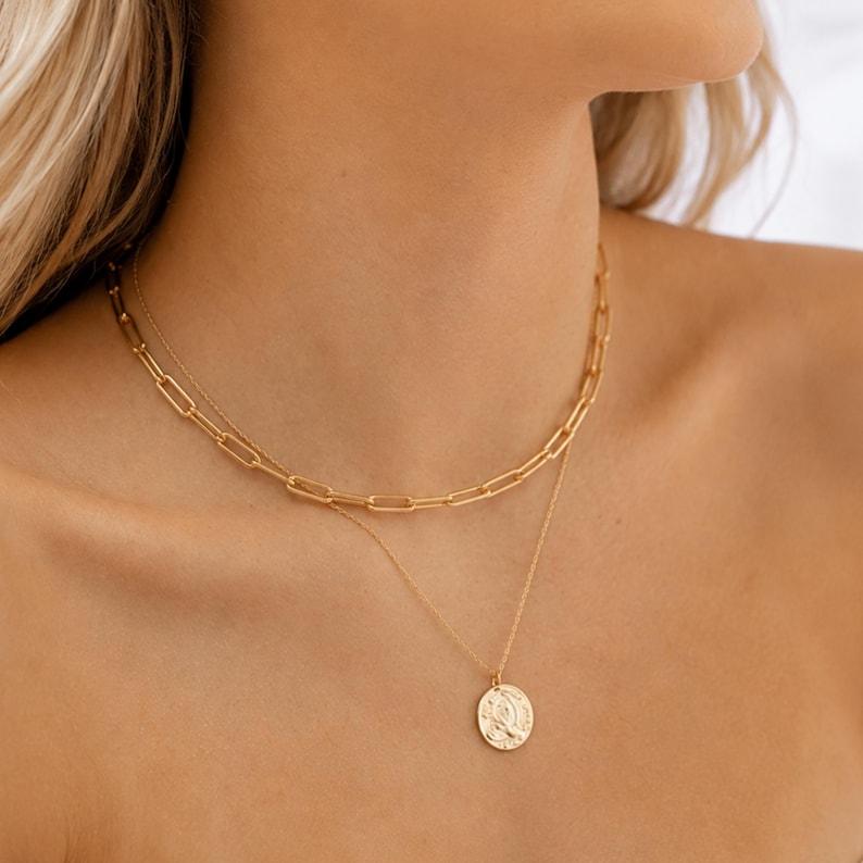 May include: A layered gold necklace set consisting of two distinct chains. The upper chain features elongated rectangular links, while the lower chain is a delicate cable style with a round, embossed coin pendant hanging from the center.
