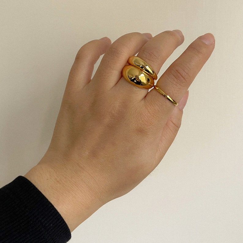 Op de afbeelding: Een set van drie goudkleurige ringen gedragen aan de vingers. Twee ringen hebben een dik, glad en afgerond koepelontwerp en zijn opgestapeld op de middelvinger, terwijl een derde, dunnere, effen gouden ring aan de ringvinger zit.