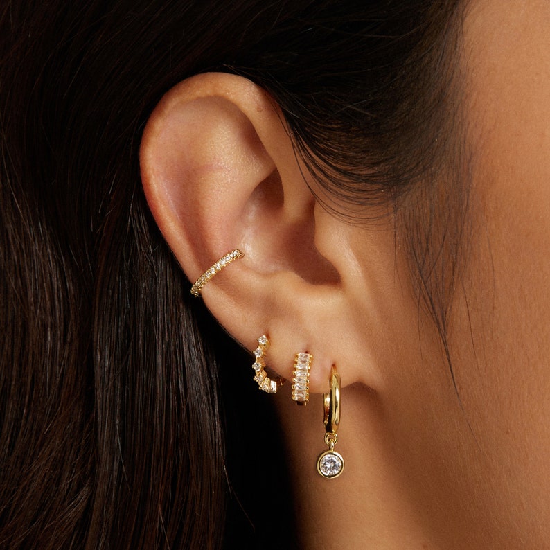 May include: A close-up of a person's ear with multiple gold earrings. The earrings are all adorned with small, clear cubic zirconia stones. The earrings include a small hoop, a larger hoop with a dangling stone, and two ear cuffs.
