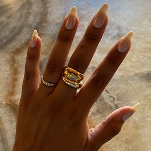 Op de afbeelding: Een goudkleurige ring met een dikke, gladde, ronde band en een tweede, dunnere ring met een getextureerde schakelketting aan dezelfde vinger. De ringen zijn gepolijst en reflecterend, met een metallic afwerking op de huid van de vinger.