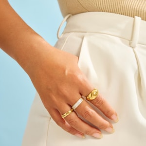 Op de afbeelding: Een set van drie goudkleurige ringen gedragen aan de vingers. E&eacute;n ring heeft een gladde, dikke, koepelvorm. De tweede ring heeft een getextureerd, glinsterend oppervlak. De derde ring is een dunne, gepolijste gouden band met een gladde afwerking.
