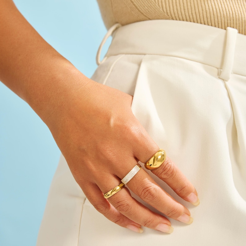 Op de afbeelding: Een set van drie goudkleurige ringen gedragen aan de vingers. E&eacute;n ring heeft een gladde, dikke, koepelvorm. De tweede ring heeft een getextureerd, glinsterend oppervlak. De derde ring is een dunne, gepolijste gouden band met een gladde afwerking.