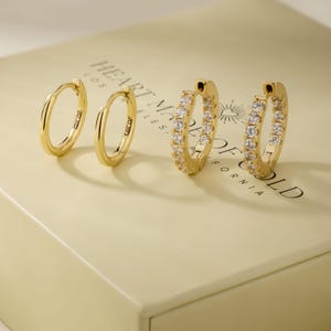 May include: Four gold hoop earrings. Two are plain, and two are encrusted with small, clear gemstones. The earrings are displayed on a light-colored surface with the text "HEART MADE OF GOLD" printed on it.