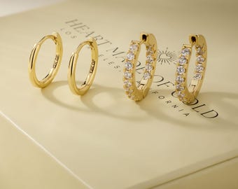May include: Four gold hoop earrings. Two are plain, and two are encrusted with small, clear gemstones. The earrings are displayed on a light-colored surface with the text "HEART MADE OF GOLD" printed on it.