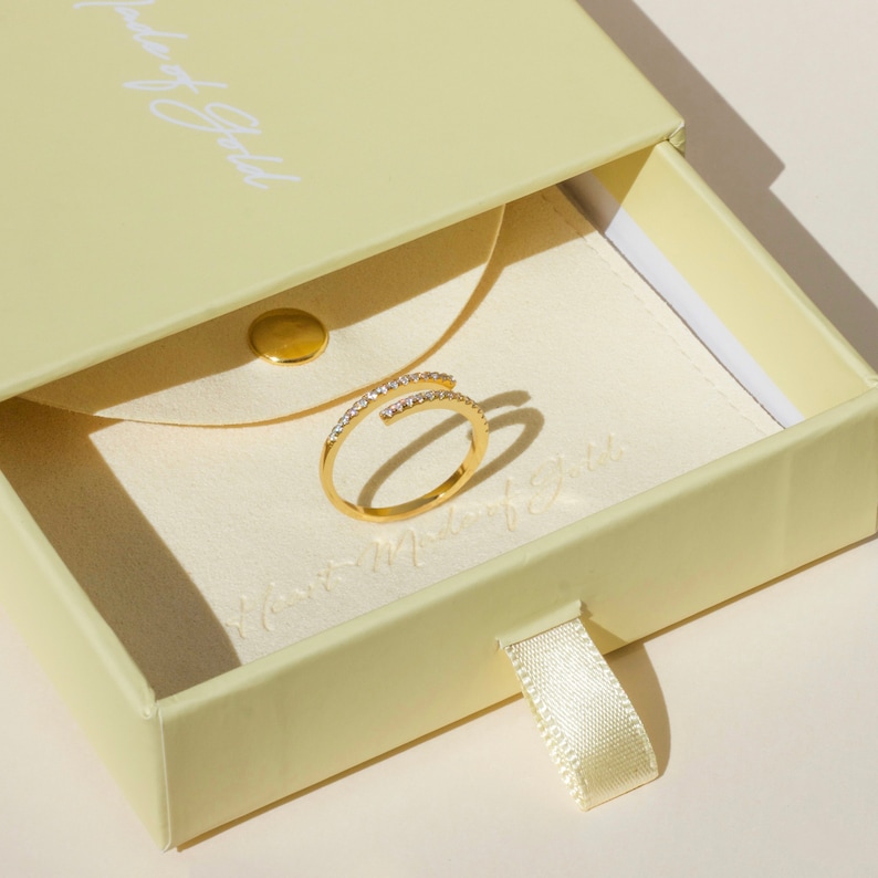 May include: A gold ring with a row of small clear stones on each side of the band. The ring is in a yellow box with a white ribbon.