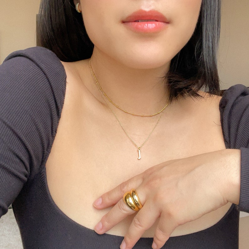 Op de afbeelding: Een gelaagde gouden kettingenset met een fijne kralenketting en een tweede ketting met een kleine letter J-hanger. Een dikke, gladde, gepolijste gouden ring wordt aan de vinger gedragen. De sieraden worden gedragen bij een zwart shirt.