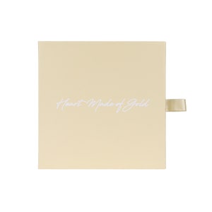 May include: A square, light beige box with the text "Heart Made of Gold" in elegant white script. A matching satin ribbon is attached to the side for opening. The box is set against a white background.