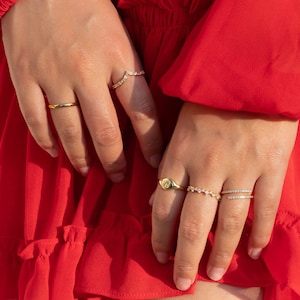 May include: A hand wearing four gold rings with clear stones. The rings are a thin band, a v-shaped ring, a ring with a single oval stone, and a ring with a row of stones.