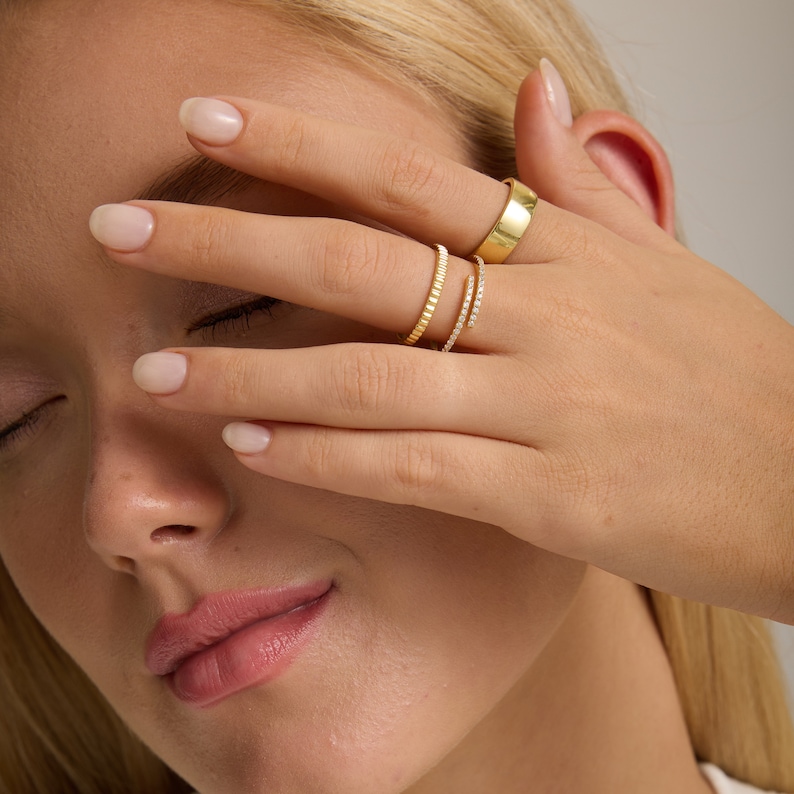 May include: A hand wearing four gold rings. The rings are stacked on the ring finger. The top ring is a thick band, the next ring is a thin band with small diamonds, the third ring is a thin band with small diamonds, and the bottom ring is a thin band with small diamonds.