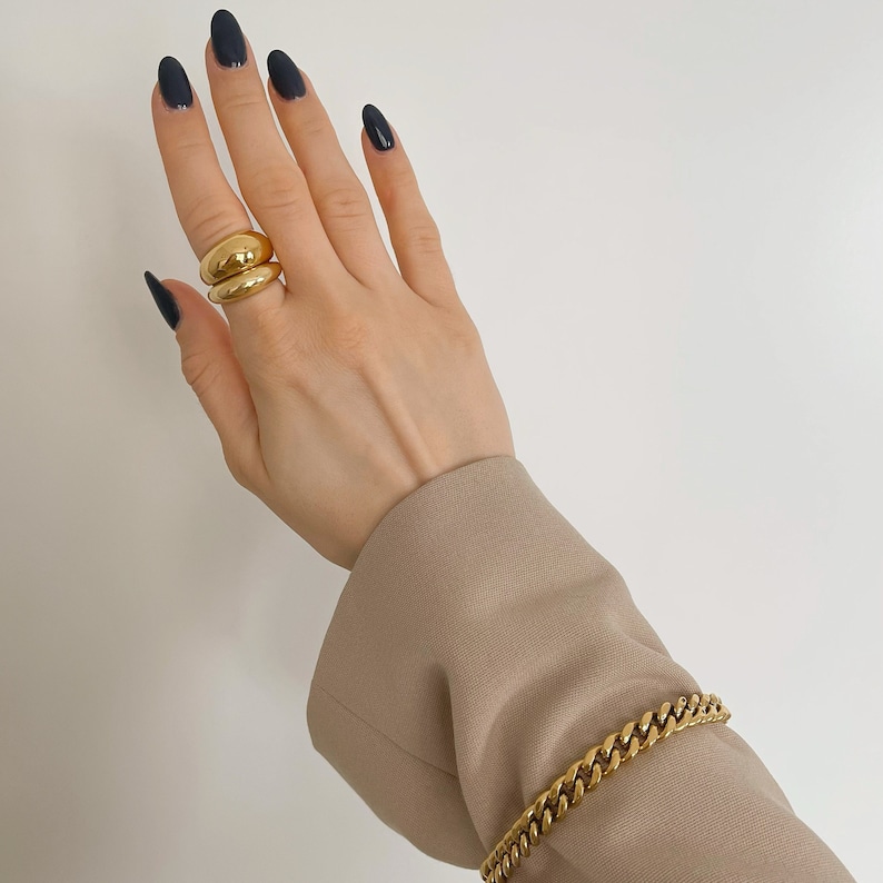 Op de afbeelding: Een goudkleurige ring met twee gestapelde, gladde en afgeronde banden gedragen aan een vinger, gecombineerd met een goudkleurige gourmetkettingarmband om de pols, zichtbaar tegen een neutrale achtergrond met een beige mouw.