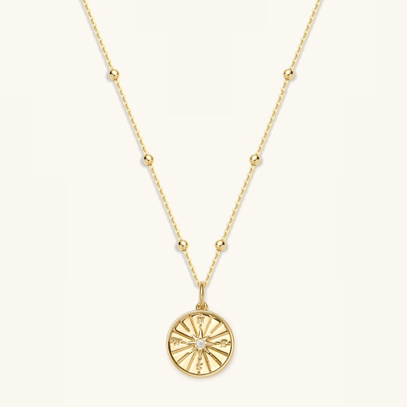 May include: A gold necklace featuring a compass pendant. The pendant has a small diamond at its centre and is surrounded by sunburst rays. The chain has small, evenly spaced gold beads.
