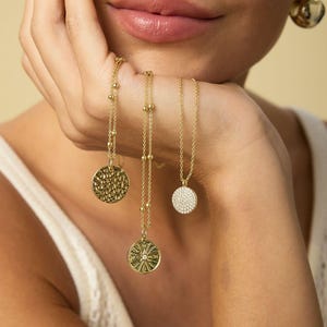 May include: Three gold necklaces with circular pendants. One pendant has a textured surface, another features a sunburst design with a central gem, and the third is covered in small, sparkling stones. The necklaces are displayed on a person's hand.