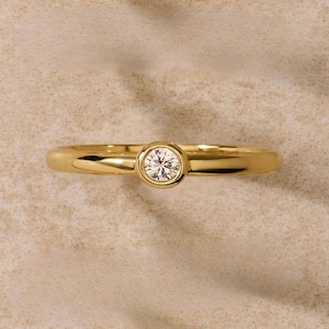 Puede incluir: Un anillo de oro con un diamante redondo y transparente en el centro. La banda es de oro pulido. El anillo se muestra sobre una superficie neutra y texturizada.