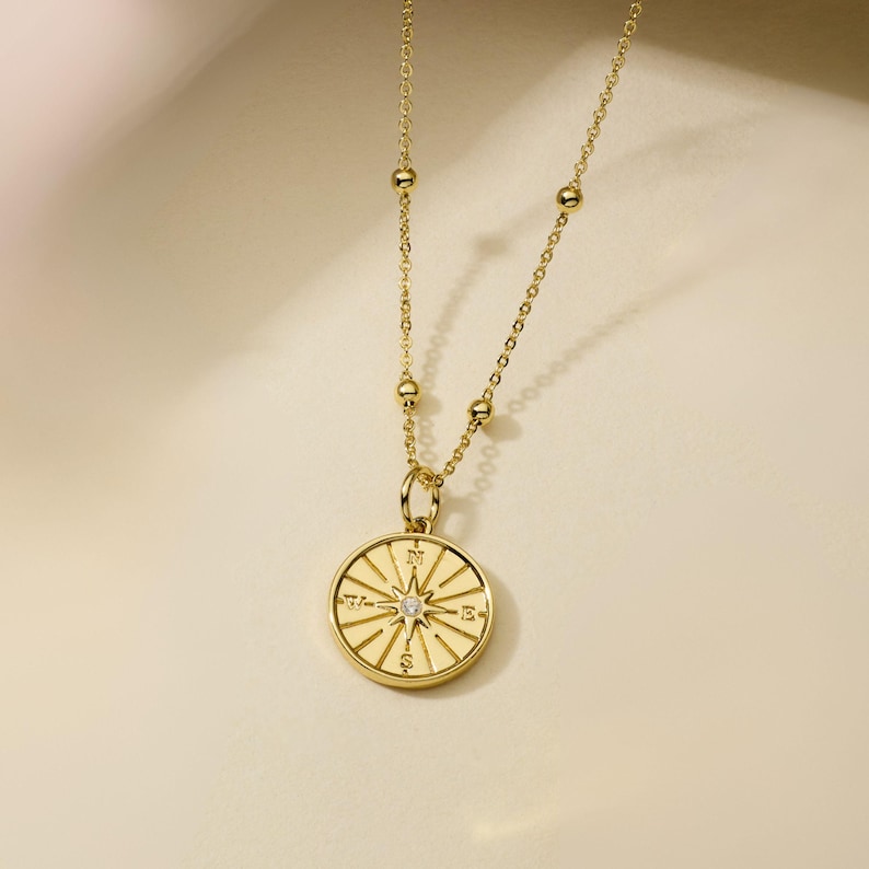 May include: A gold necklace featuring a compass pendant. The pendant has a starburst design with a small, clear gemstone at the centre. The chain has small, round beads along its length. The necklace is set against a neutral background.