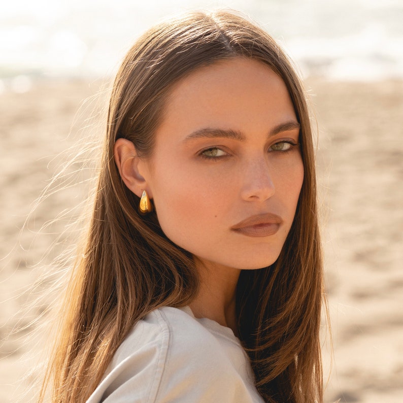 May include: A woman with long brown hair wearing a gold hoop earring. The earring is a simple, smooth, teardrop shape.