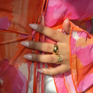 Op de afbeelding: Een doorschijnende mesh top met een abstracte oranje en roze aquarelprint en een striksluiting aan de voorzijde. Aan de vingers zitten twee gouden ringen: een gladde, brede koepelvormige ring en een gestructureerde ring met een kleine steen.