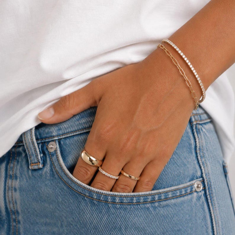 Op de afbeelding: Een goudkleurige sieradenset bestaande uit twee armbanden en drie ringen. De ene armband heeft een delicaat schakelontwerp, terwijl de andere een dunne band is ingelegd met kleine heldere steentjes. De ringen omvatten een gladde bolle ring en twee dunne ringen.