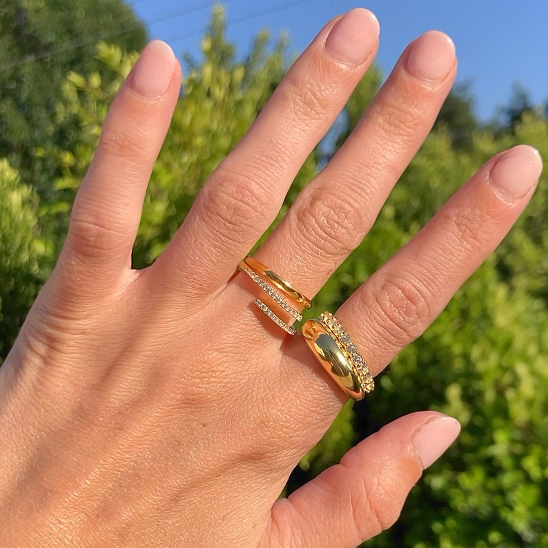May include: Two gold rings with clear stones on a hand. The ring on the index finger is a thin band with a single row of stones. The ring on the middle finger is a thicker band with multiple rows of stones.