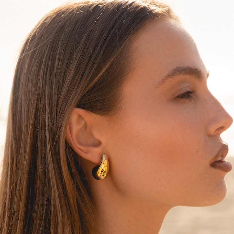 May include: A close-up of a person wearing a pair of gold hoop earrings. The earrings are large and have a smooth, rounded shape.