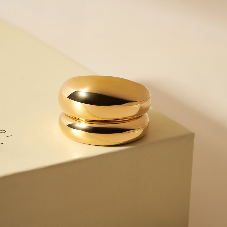 Op de afbeelding: Een gepolijste gouden ring met een dik, afgerond en glad bandontwerp. Het metalen oppervlak is sterk reflecterend en vertoont heldere accenten en diepe schaduwen die de gebogen, dikke en doorlopende cirkelvormige structuur benadrukken.