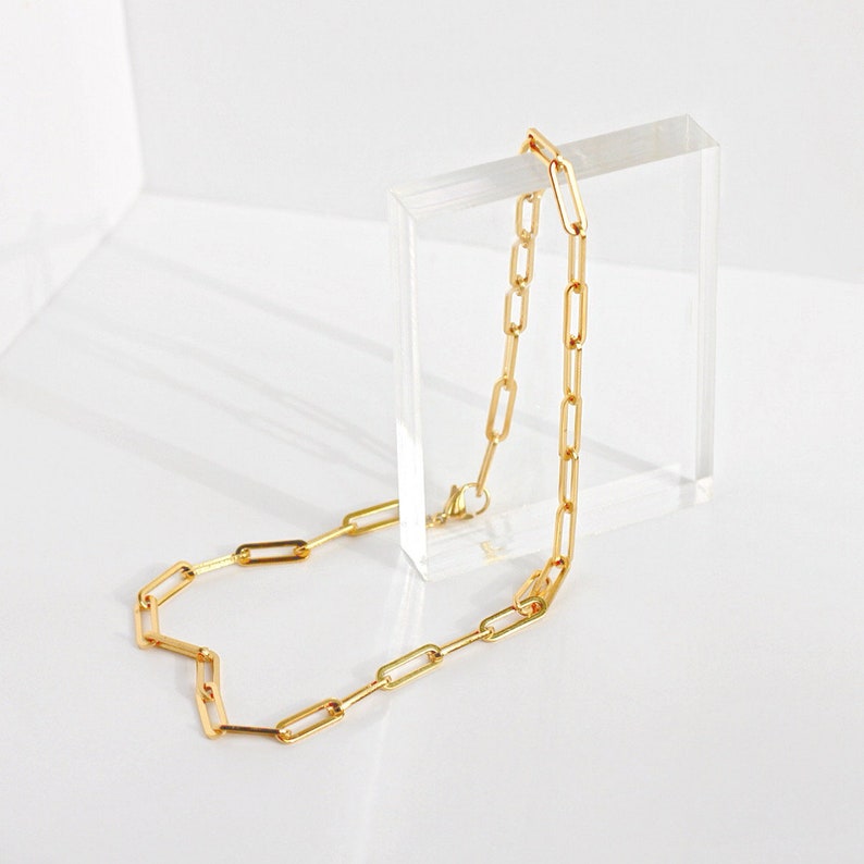 May include: A gold-toned chain necklace featuring elongated, rectangular paperclip-style links. The necklace is draped over a clear, rectangular acrylic block, showcasing the uniform metallic links and a small lobster claw clasp closure.